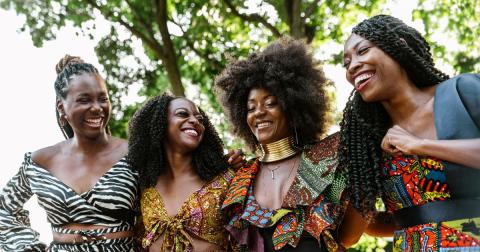 Four black women standing shoulder to shoulder smiling at each other. One Black woman with braids wearing a zebra print two piece dress. Three women wearing colorful African print outfits.  