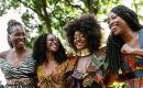 Four black women standing shoulder to shoulder smiling at each other. One Black woman with braids wearing a zebra print two piece dress. Three women wearing colorful African print outfits.  