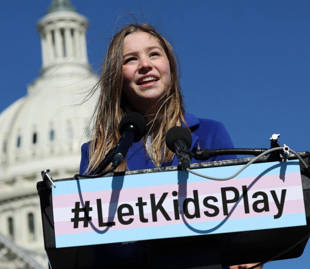 A white girl standing at a podium that says #LetKidsPlay with a blue sky capitol dome in the background